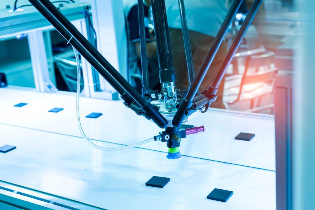 Close-up of a precision delta-style robotic arm in a medical device manufacturing clean room, positioning components on an illuminated surface. The robotic system features multiple black carbon fiber struts converging to a blue suction gripper, demonstrating advanced automation technology used in FDA-regulated medical device production environments where cybersecurity safeguards are essential throughout the manufacturing process and medical device cybersecurity.