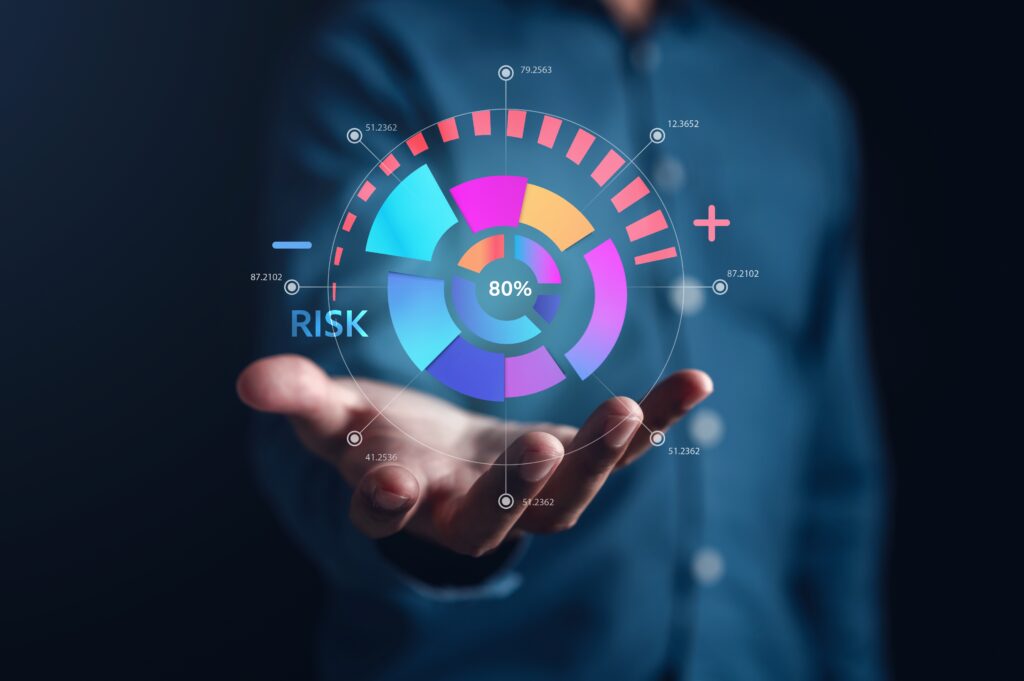 A business professional holds out their hand, displaying a digital, holographic risk assessment interface floating above their palm. The circular visualization shows an 80% risk indicator in the center, surrounded by colorful segmented rings in blue, pink, purple, and orange. Data points are marked around the perimeter with numerical values like 87.2102, 79.2563, and 51.2362. The word 'RISK' appears prominently in blue on the left side, supporting the idea healthcare risk assessment with a minus symbol, while a plus symbol appears on the right. The background is dark blue, creating contrast with the vibrant data visualization.