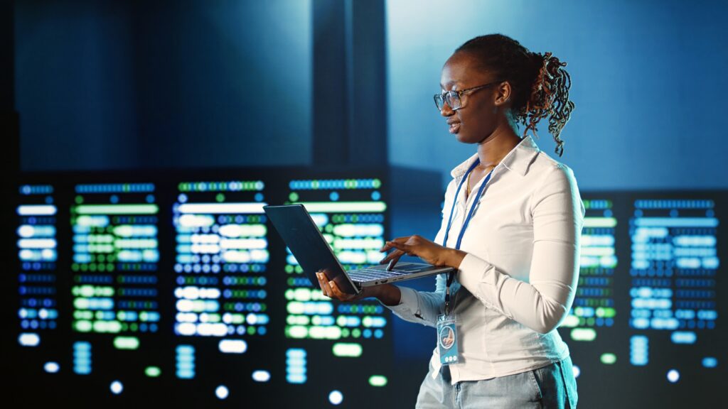 Cybersecurity analyst working in a security operations center (SOC), monitoring systems on a laptop against data center background.”